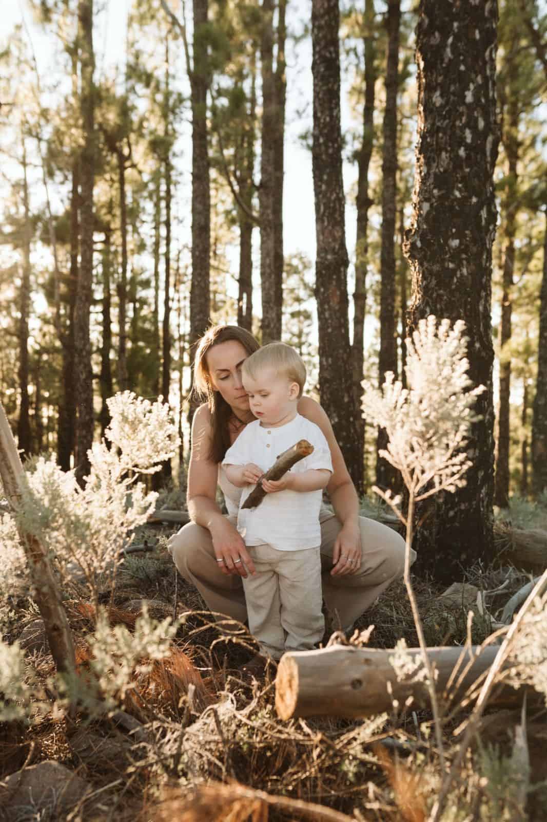 Niño pequeño jugando en la naturaleza sujetando un tronco de madera y acompañado por su madre