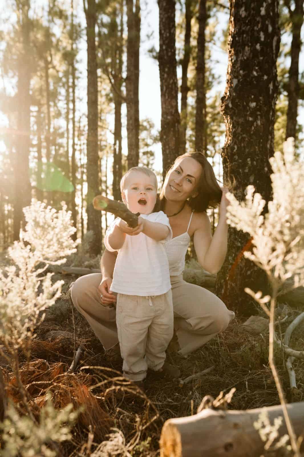 Niño pequeño jugando en la naturaleza sujetando un tronco de madera y acompañado por su madre