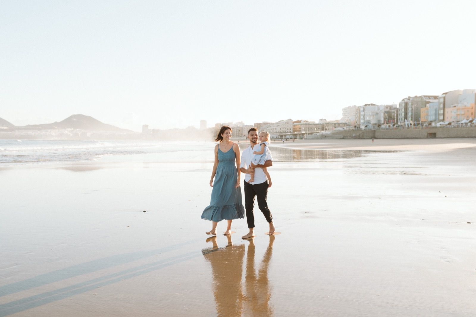 Familia formada por hombre, mujer y niña pequeña posando a la cámara sonriendo y caminando por la orilla de la Playa de Las Canteras en Gran Canaria durante las horas de marea baja. Fotografía realizada por el fotógrafo de familias en Gran Canaria Héctor Cardeñosa.