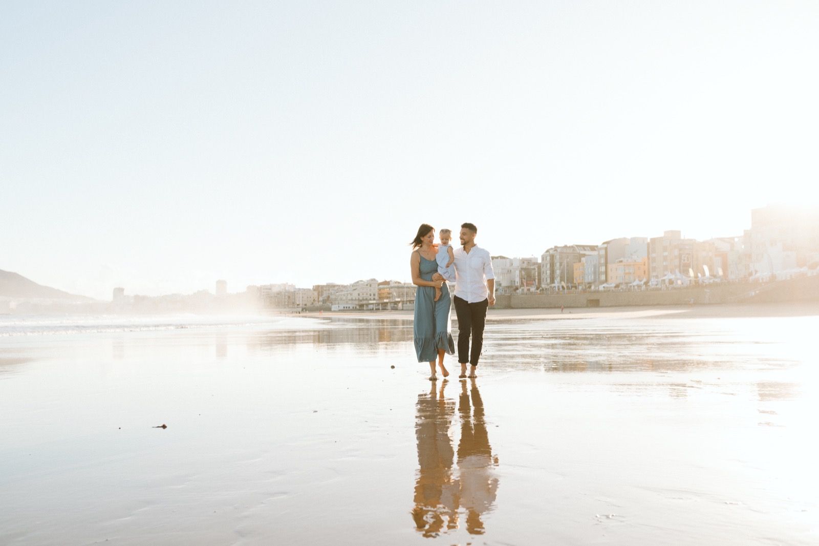 Familia formada por hombre, mujer y niña pequeña posando a la cámara sonriendo y caminando por la orilla de la Playa de Las Canteras en Gran Canaria durante las horas de marea baja. Fotografía realizada por el fotógrafo de familias en Gran Canaria Héctor Cardeñosa.