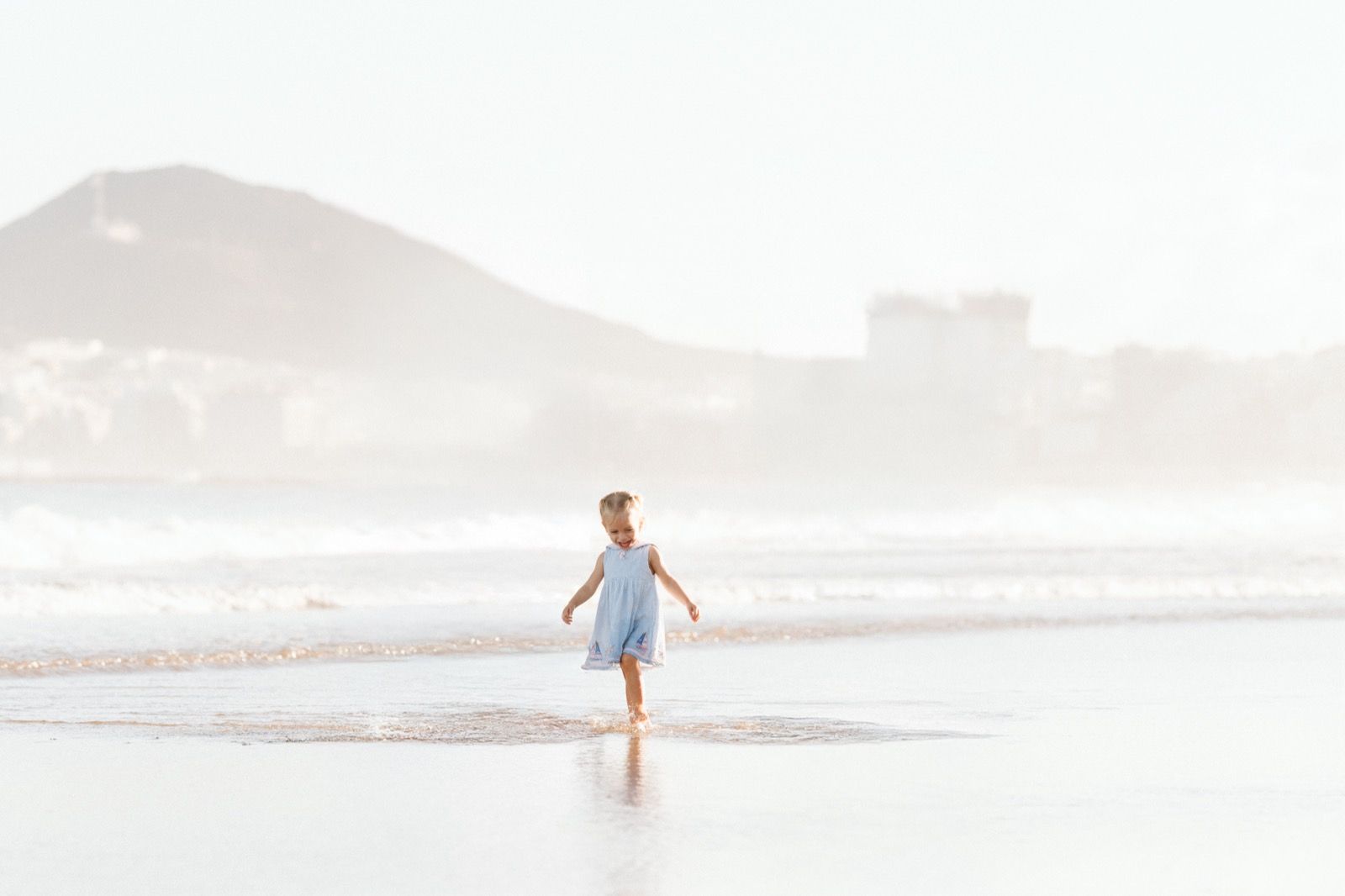 Niña con vestido azul corriendo en la orilla de la playa de Las Canteras durante la mañana en marea baja. Fotografía realizada por el fotógrafo de familias en Gran Canaria Héctor Cardeñosa