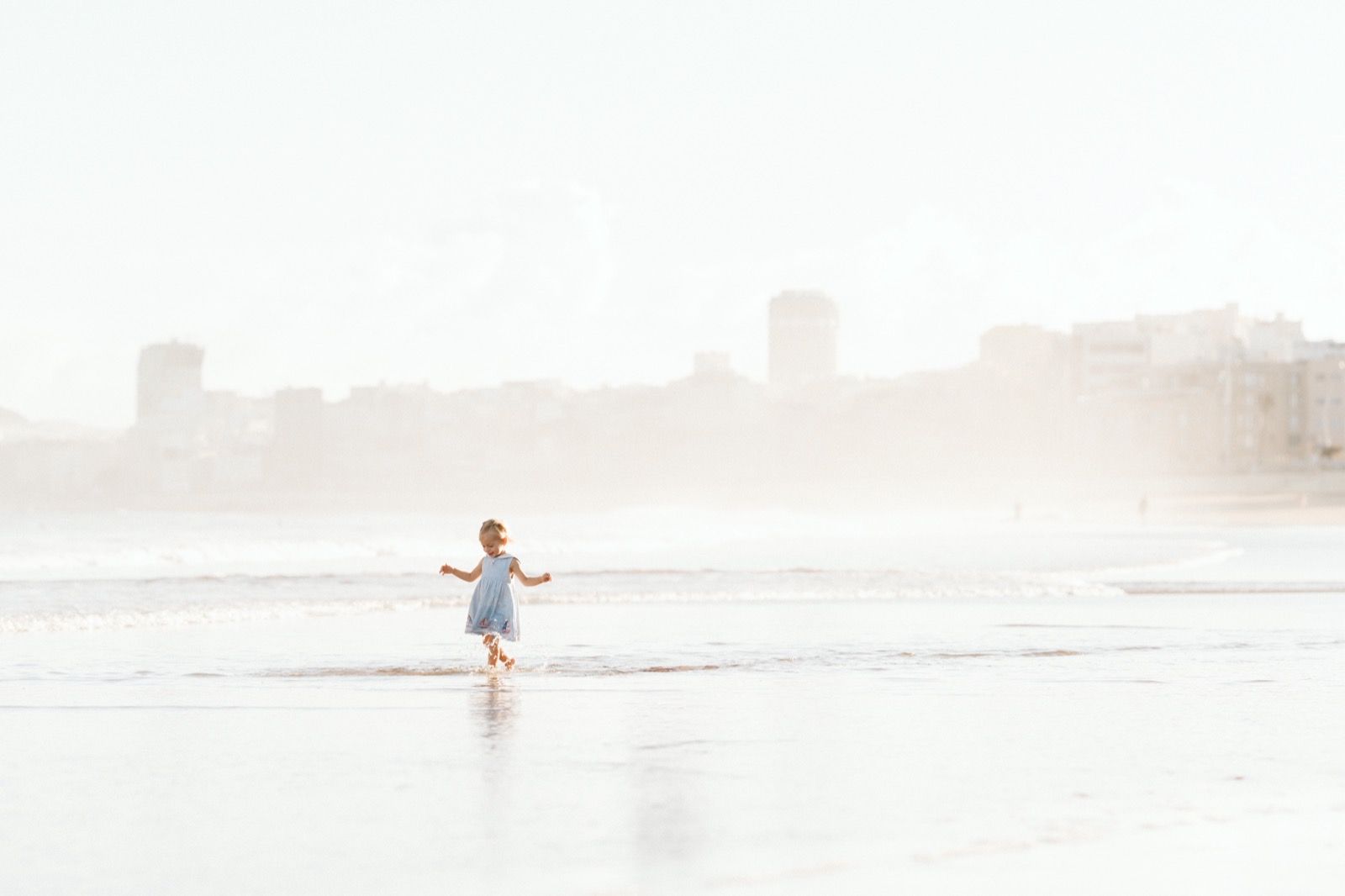 Niña con vestido azul corriendo en la orilla de la playa de Las Canteras durante la mañana en marea baja. Fotografía realizada por el fotógrafo de familias en Gran Canaria Héctor Cardeñosa