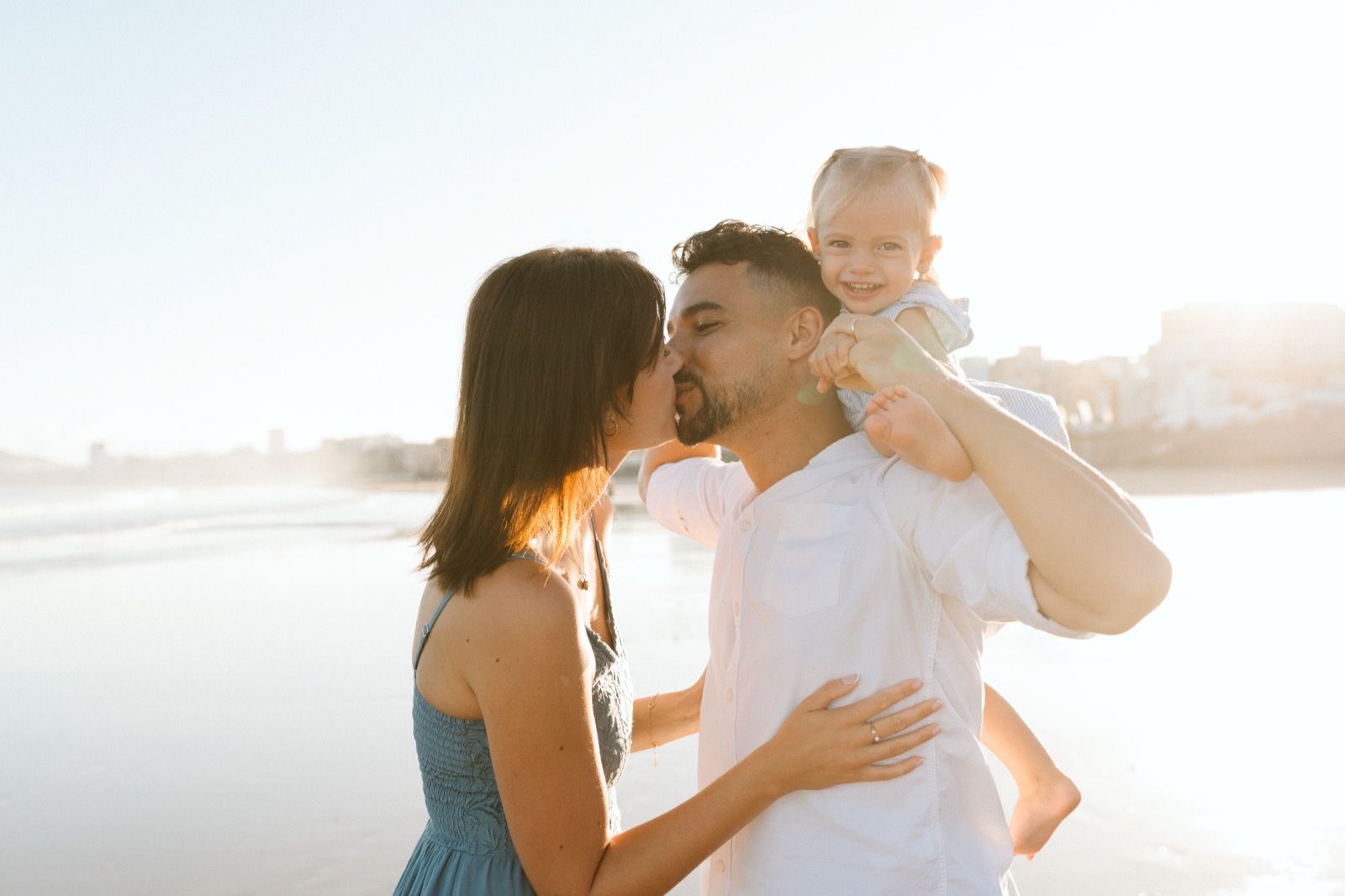 Familia formada por hombre, mujer y niña pequeña posando a la cámara sonriendo y caminando por la orilla de la Playa de Las Canteras en Gran Canaria durante las horas de marea baja. Fotografía realizada por el fotógrafo de familias en Gran Canaria Héctor Cardeñosa.