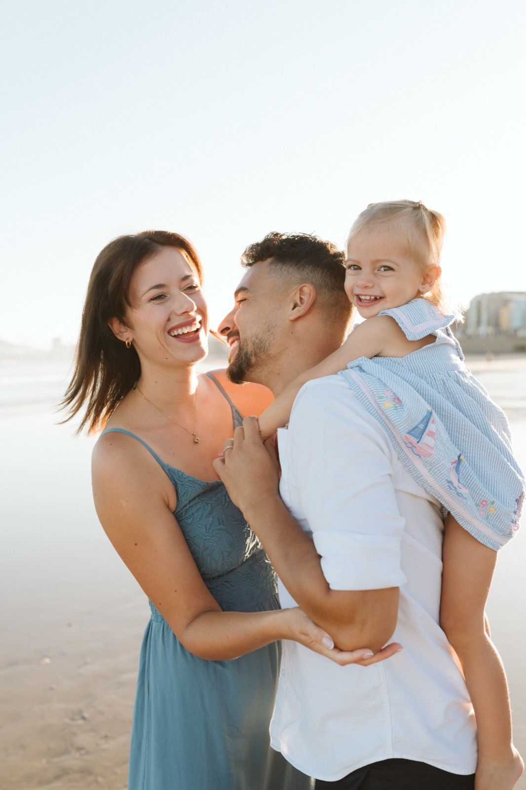 Familia formada por hombre, mujer y niña pequeña posando a la cámara sonriendo y caminando por la orilla de la Playa de Las Canteras en Gran Canaria durante las horas de marea baja. Fotografía realizada por el fotógrafo de familias en Gran Canaria Héctor Cardeñosa.