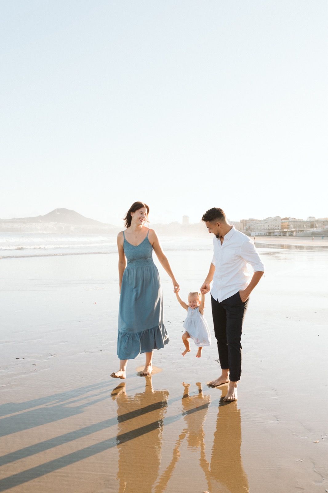 Familia formada por hombre, mujer y niña pequeña posando a la cámara sonriendo y caminando por la orilla de la Playa de Las Canteras en Gran Canaria durante las horas de marea baja. Fotografía realizada por el fotógrafo de familias en Gran Canaria Héctor Cardeñosa.