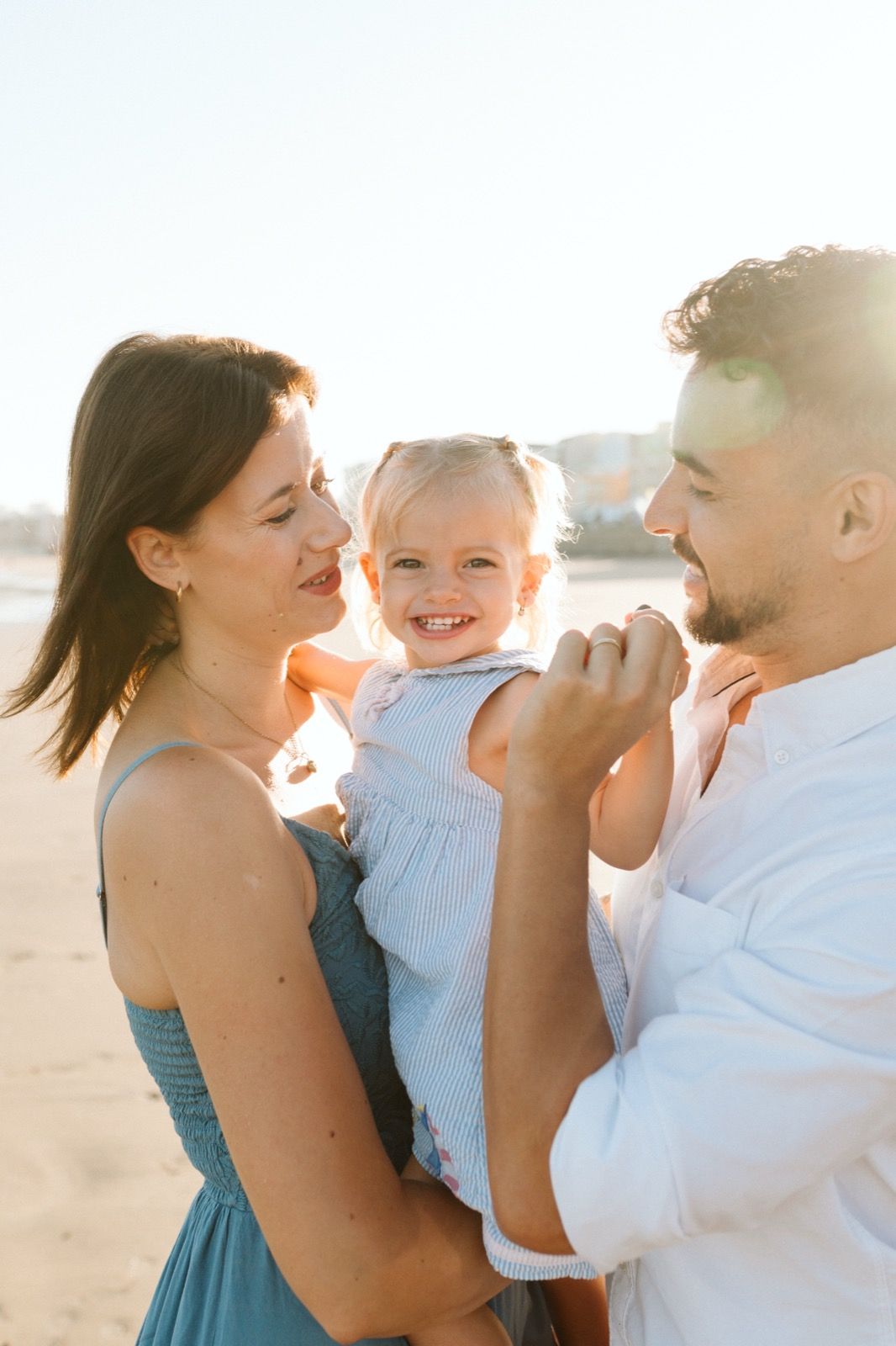 Familia formada por hombre, mujer y niña pequeña posando a la cámara sonriendo y caminando por la orilla de la Playa de Las Canteras en Gran Canaria durante las horas de marea baja. Fotografía realizada por el fotógrafo de familias en Gran Canaria Héctor Cardeñosa.