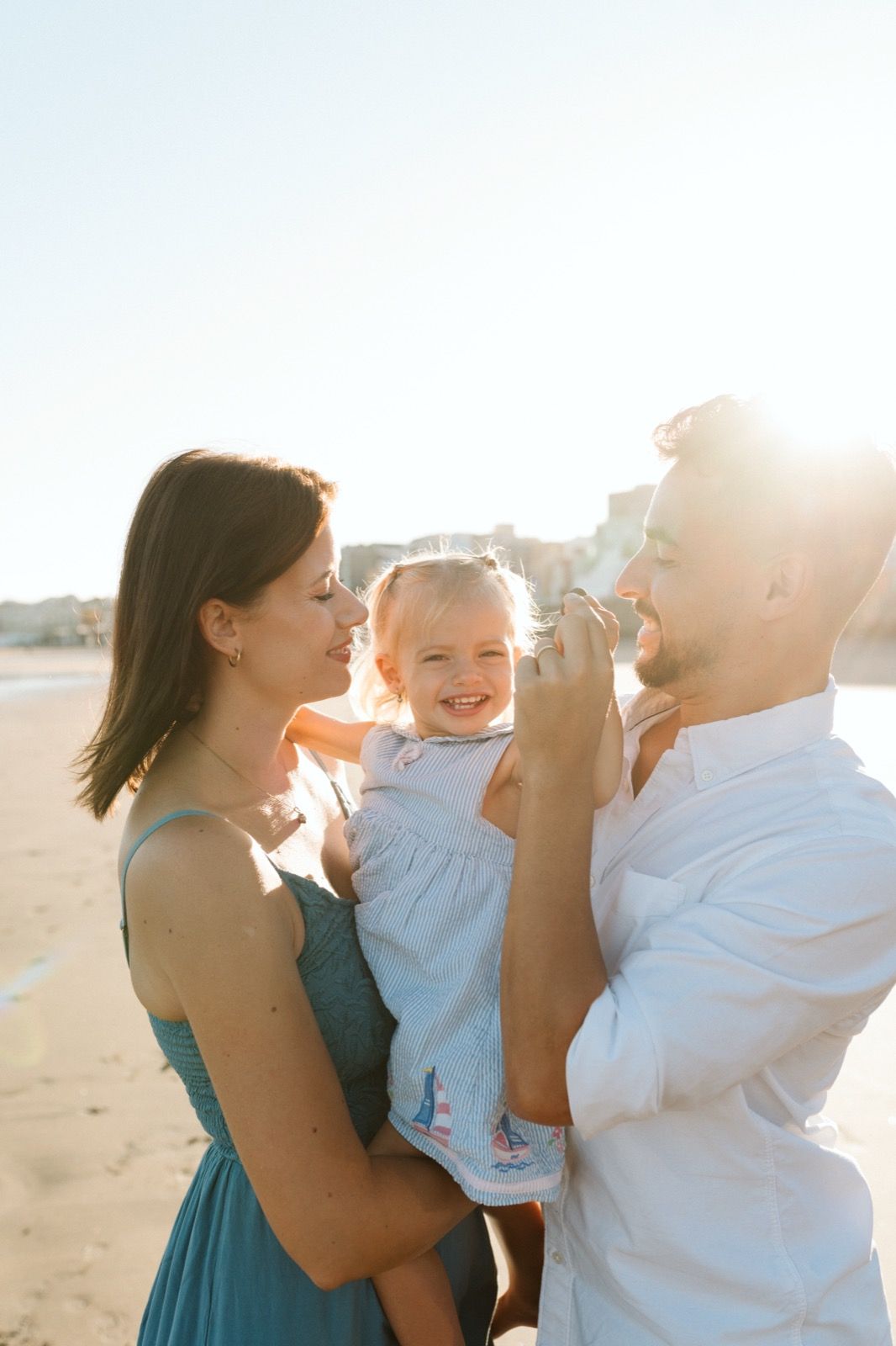 Familia formada por hombre, mujer y niña pequeña posando a la cámara sonriendo y caminando por la orilla de la Playa de Las Canteras en Gran Canaria durante las horas de marea baja. Fotografía realizada por el fotógrafo de familias en Gran Canaria Héctor Cardeñosa.
