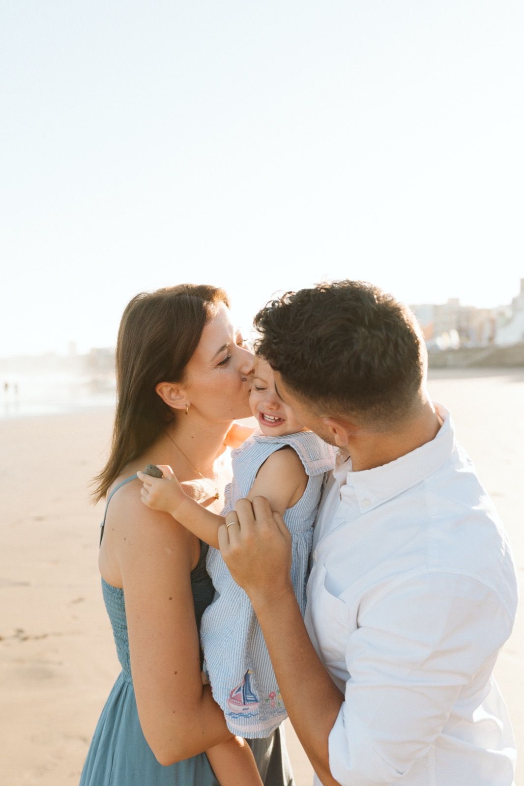 Familia formada por hombre, mujer y niña pequeña posando a la cámara sonriendo y besando a su hija por la orilla de la Playa de Las Canteras en Gran Canaria durante las horas de marea baja. Fotografía realizada por el fotógrafo de familias en Gran Canaria Héctor Cardeñosa.
