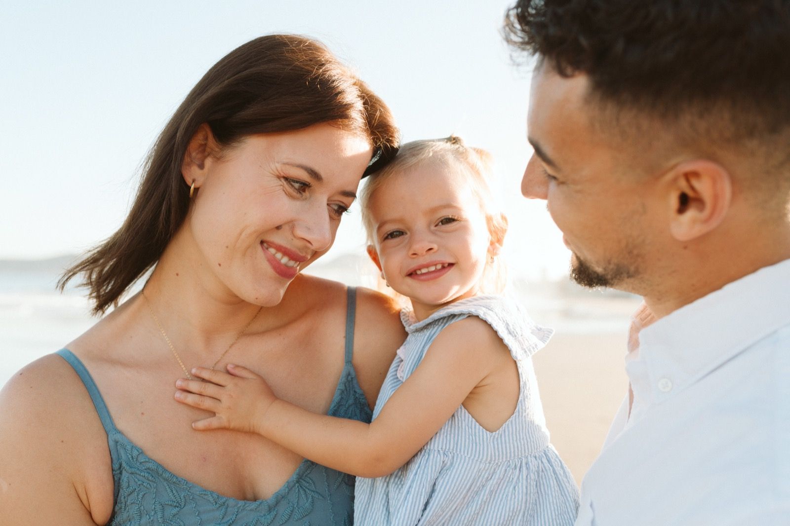 Familia formada por hombre, mujer y niña pequeña posando a la cámara sonriendo y caminando por la orilla de la Playa de Las Canteras en Gran Canaria durante las horas de marea baja. Fotografía realizada por el fotógrafo de familias en Gran Canaria Héctor Cardeñosa.