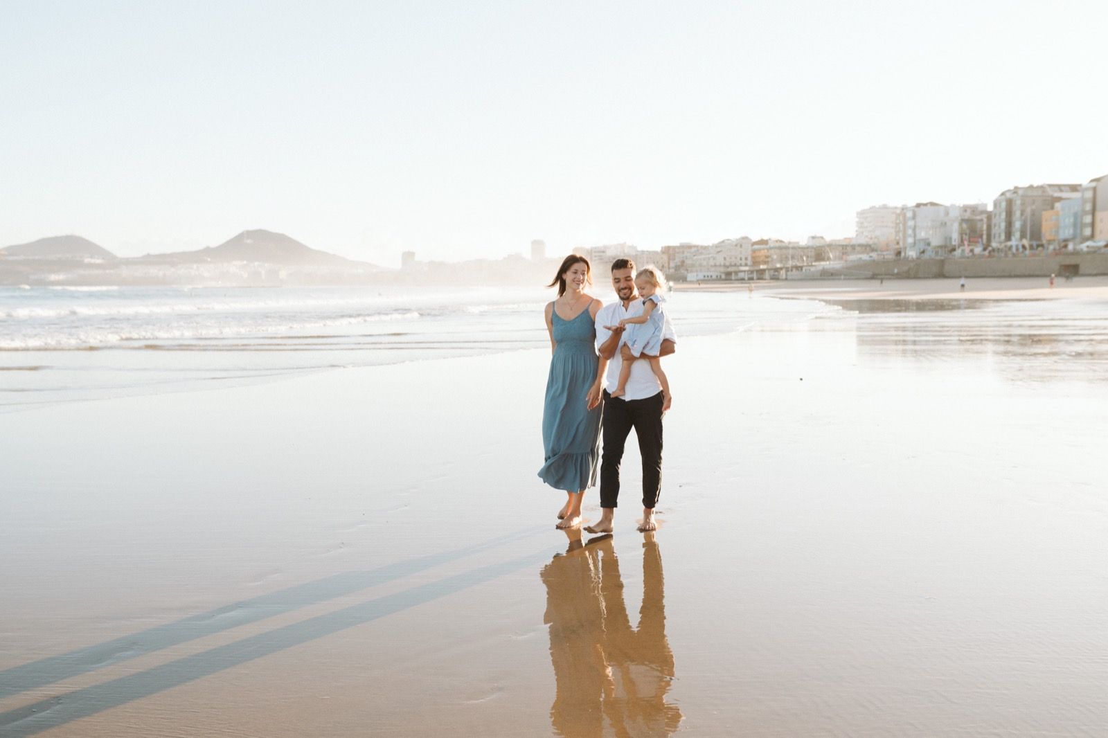 Familia formada por hombre, mujer y niña pequeña posando a la cámara sonriendo y caminando por la orilla de la Playa de Las Canteras en Gran Canaria durante las horas de marea baja. Fotografía realizada por el fotógrafo de familias en Gran Canaria Héctor Cardeñosa.