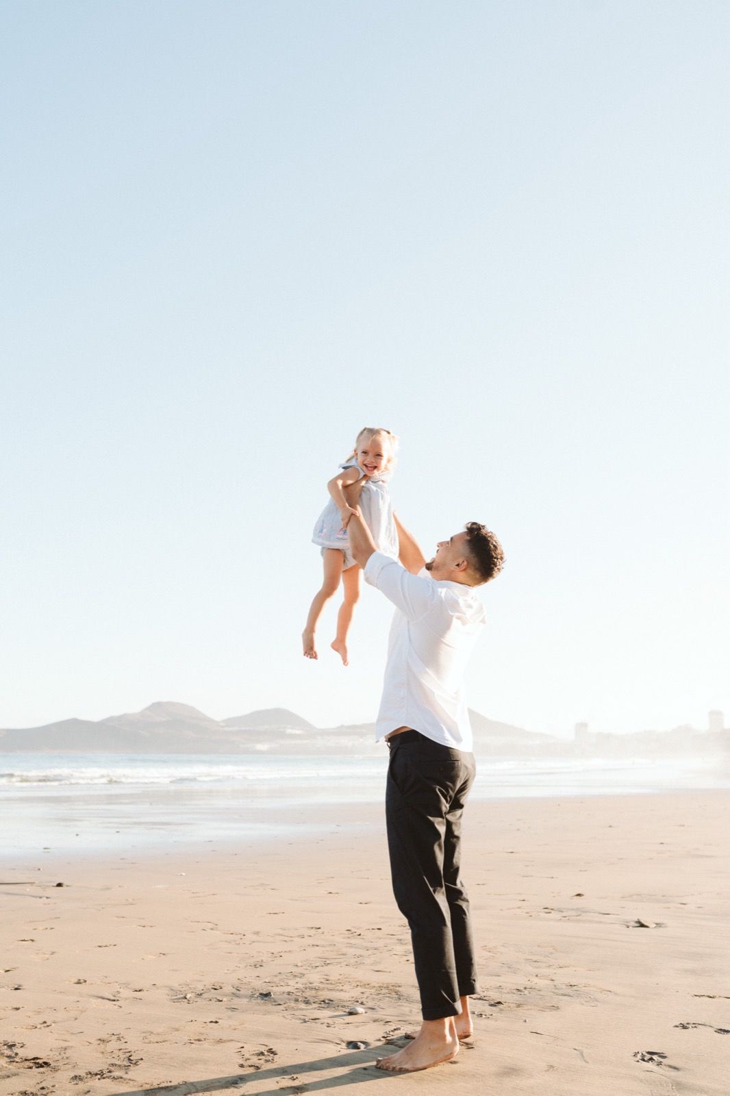 Familia formada por hombre y niña pequeña. Padre juega con su hija lanzándola al aire, ambos sonriendo en la orilla de la Playa de Las Canteras en Gran Canaria durante las horas de marea baja. Fotografía realizada por el fotógrafo de familias en Gran Canaria Héctor Cardeñosa.