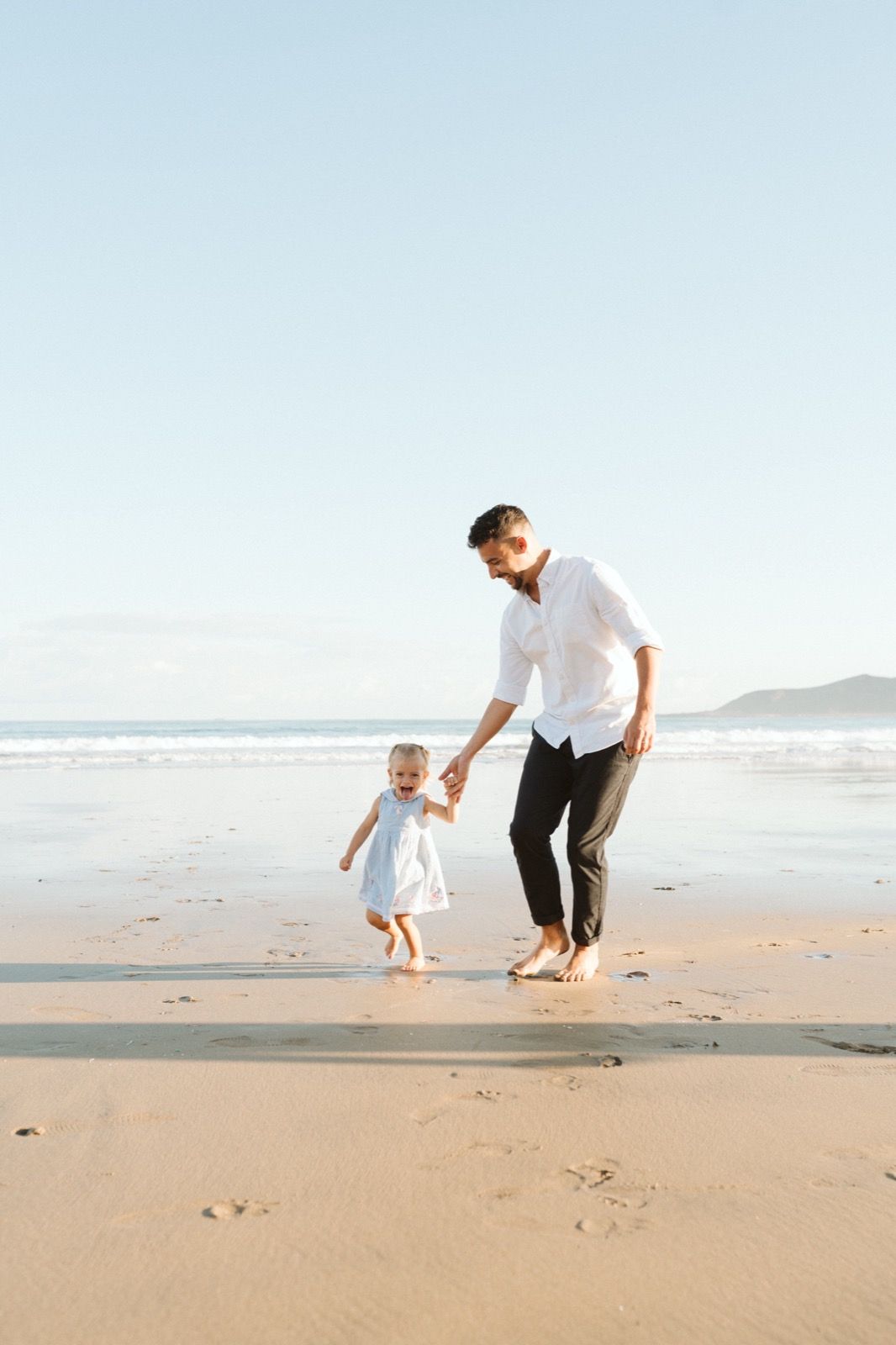 Familia formada por hombre y niña pequeña posando a la cámara sonriendo y caminando por la orilla de la Playa de Las Canteras en Gran Canaria durante las horas de marea baja. Fotografía realizada por el fotógrafo de familias en Gran Canaria Héctor Cardeñosa.