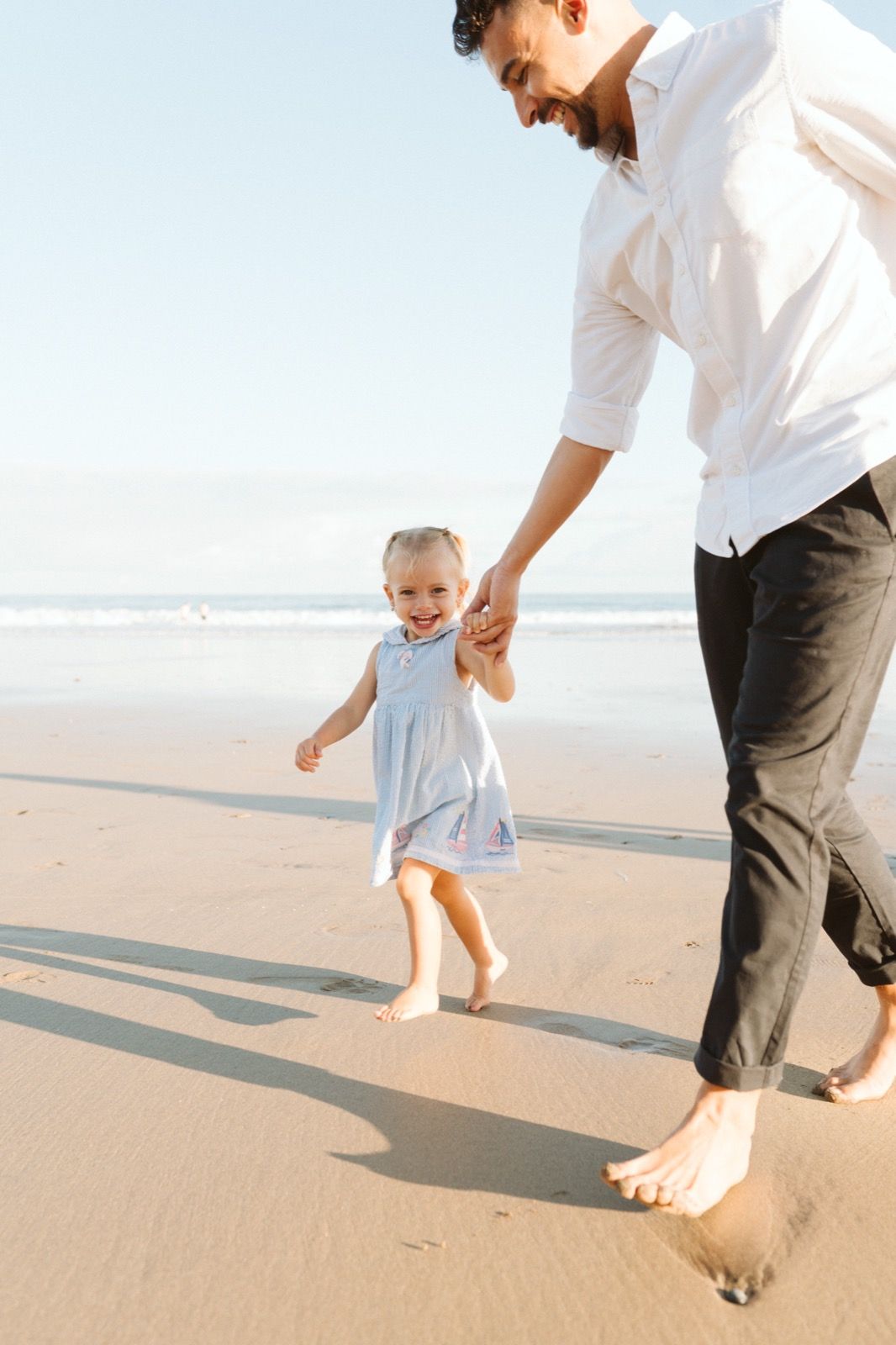 Familia formada por hombre y niña pequeña posando a la cámara sonriendo y caminando por la orilla de la Playa de Las Canteras en Gran Canaria durante las horas de marea baja. Fotografía realizada por el fotógrafo de familias en Gran Canaria Héctor Cardeñosa.
