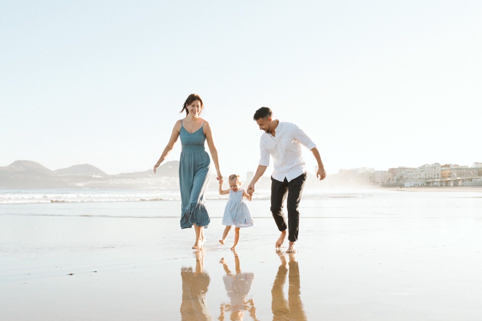 Familia formada por hombre, mujer y niña pequeña posando a la cámara sonriendo y caminando por la orilla de la Playa de Las Canteras en Gran Canaria durante las horas de marea baja. Fotografía realizada por el fotógrafo de familias en Gran Canaria Héctor Cardeñosa.