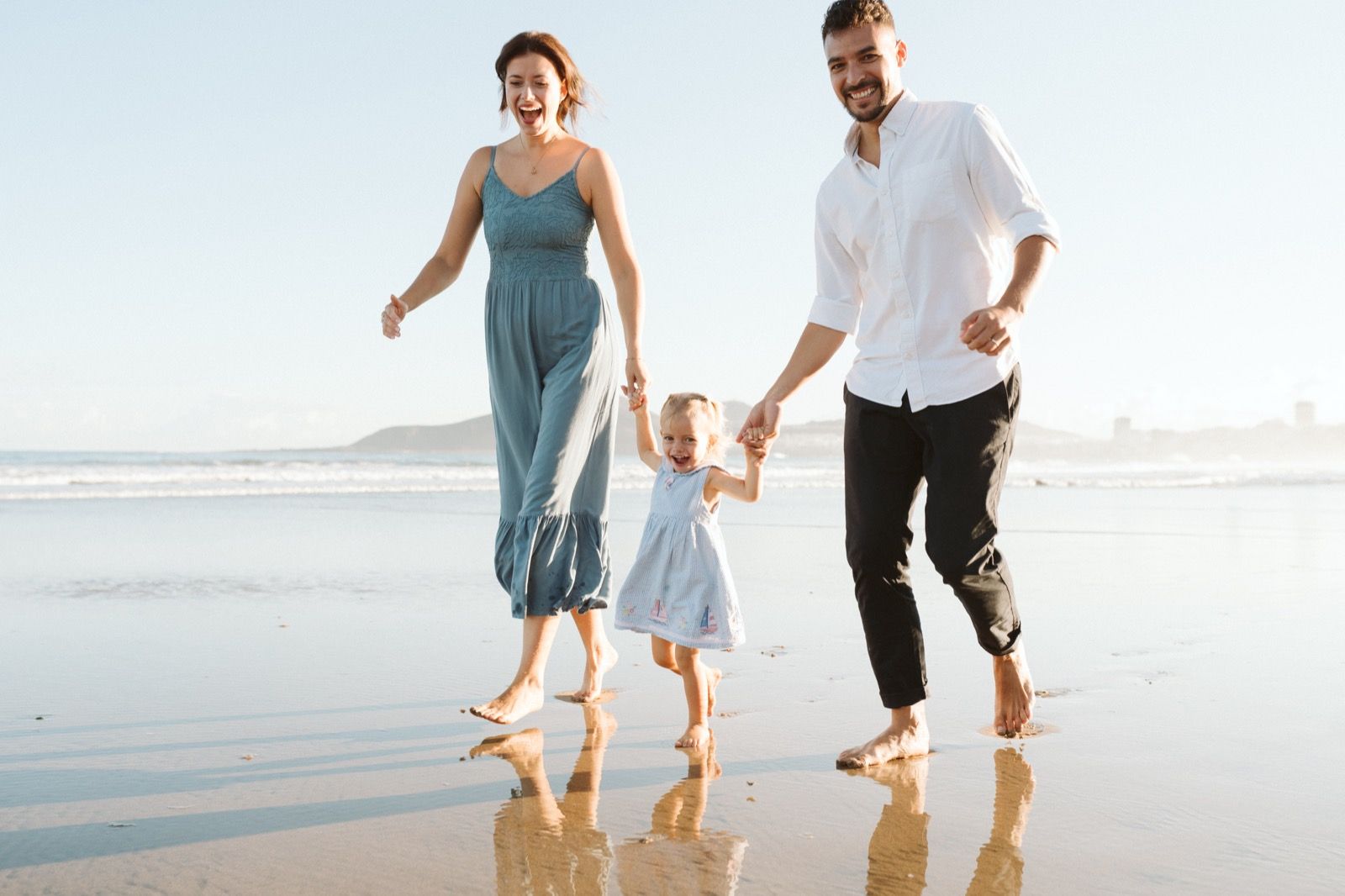 Familia formada por hombre, mujer y niña pequeña posando a la cámara sonriendo y caminando por la orilla de la Playa de Las Canteras en Gran Canaria durante las horas de marea baja. Fotografía realizada por el fotógrafo de familias en Gran Canaria Héctor Cardeñosa.