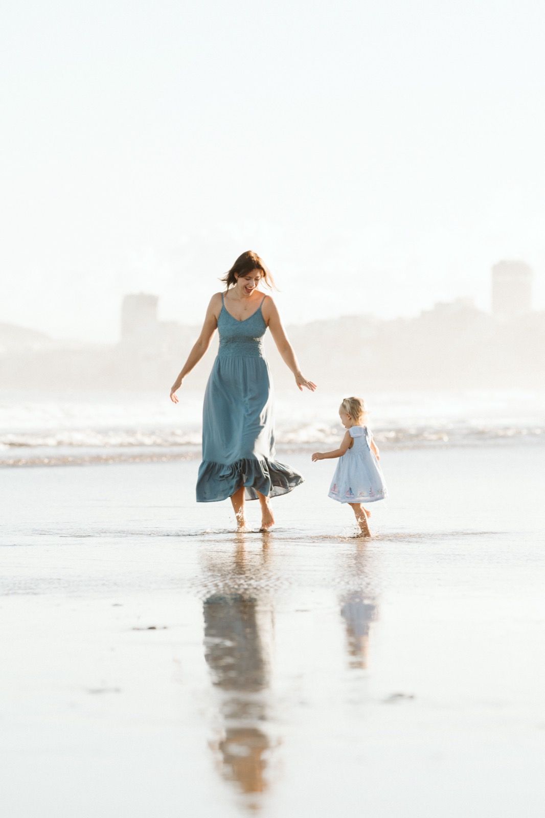 Familia formada por mujer y niña pequeña posando a la cámara sonriendo y caminando por la orilla de la Playa de Las Canteras en Gran Canaria durante las horas de marea baja. Fotografía realizada por el fotógrafo de familias en Gran Canaria Héctor Cardeñosa.
