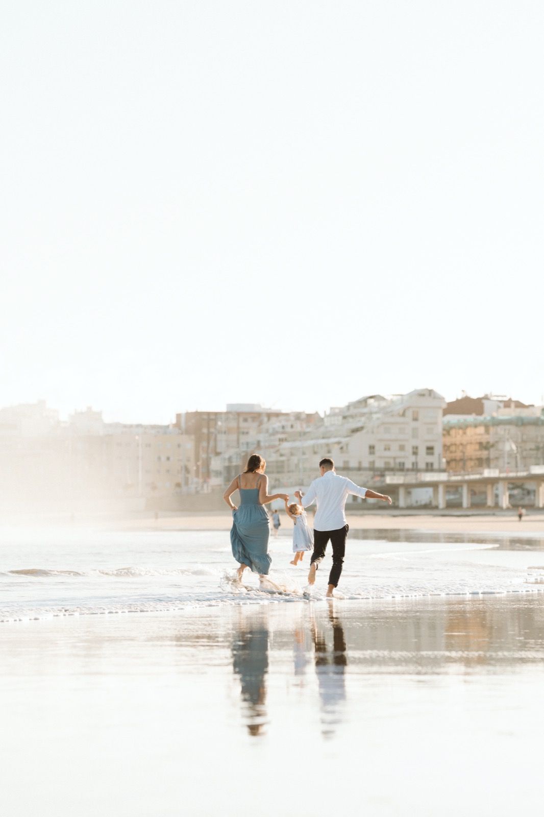 Familia formada por hombre, mujer y niña pequeña posando a la cámara sonriendo y caminando por la orilla de la Playa de Las Canteras en Gran Canaria durante las horas de marea baja. Fotografía realizada por el fotógrafo de familias en Gran Canaria Héctor Cardeñosa.
