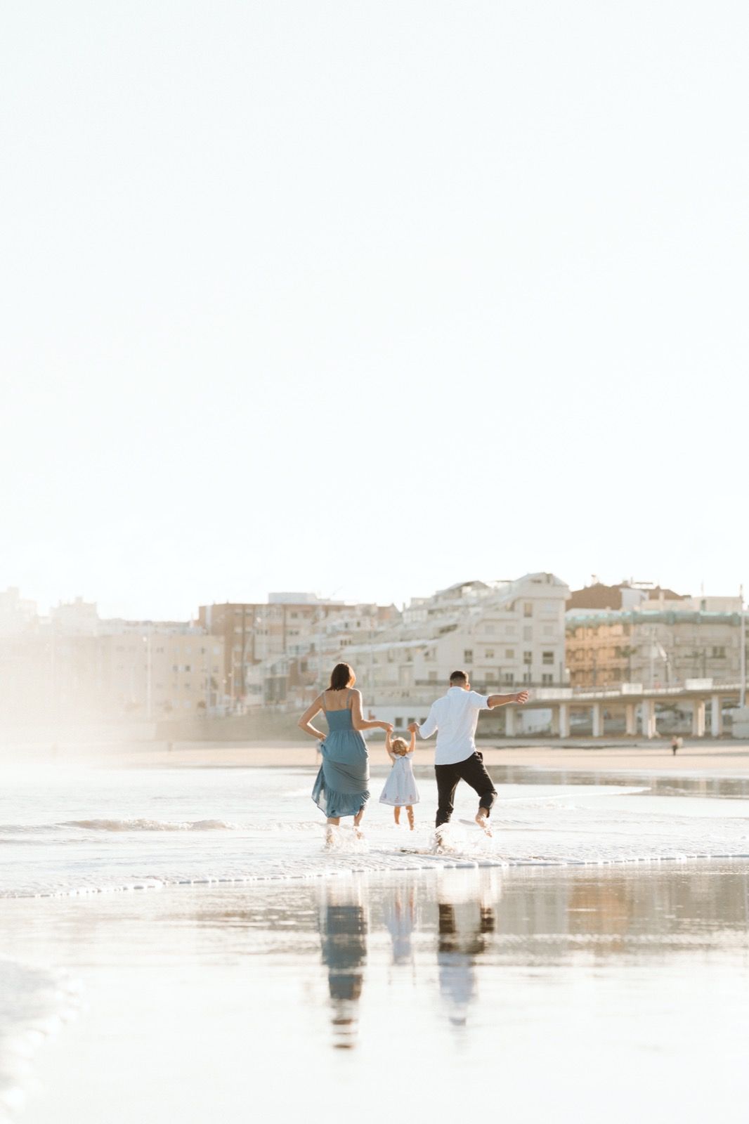 Familia formada por hombre, mujer y niña pequeña posando a la cámara sonriendo y caminando por la orilla de la Playa de Las Canteras en Gran Canaria durante las horas de marea baja. Fotografía realizada por el fotógrafo de familias en Gran Canaria Héctor Cardeñosa.