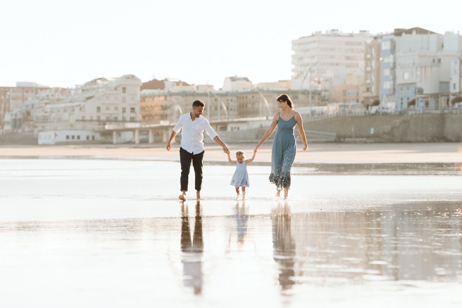 Familia formada por hombre, mujer y niña pequeña posando a la cámara sonriendo y caminando por la orilla de la Playa de Las Canteras en Gran Canaria durante las horas de marea baja. Fotografía realizada por el fotógrafo de familias en Gran Canaria Héctor Cardeñosa.
