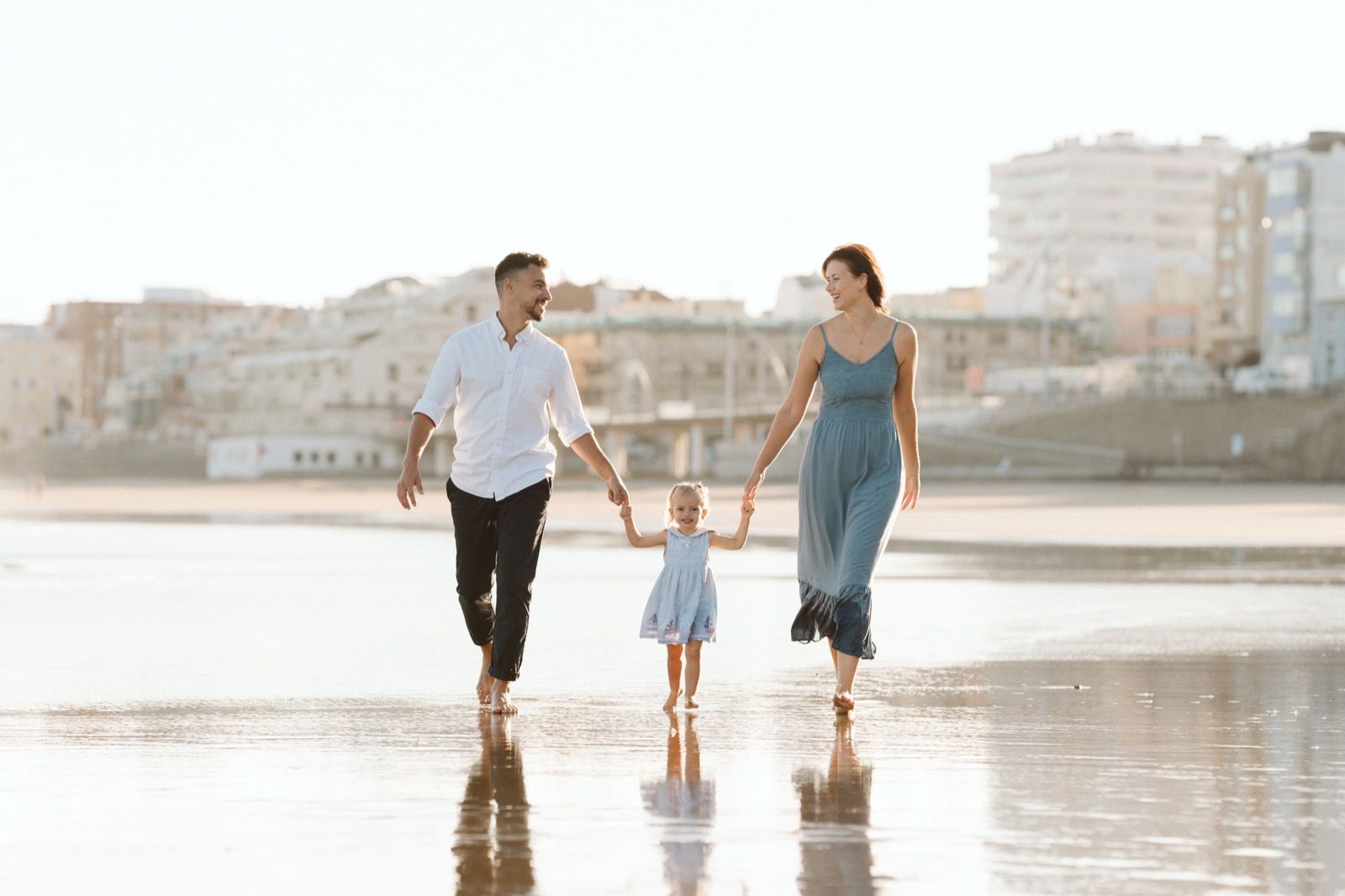 Familia formada por hombre, mujer y niña pequeña posando a la cámara sonriendo y caminando por la orilla de la Playa de Las Canteras en Gran Canaria durante las horas de marea baja. Fotografía realizada por el fotógrafo de familias en Gran Canaria Héctor Cardeñosa.
