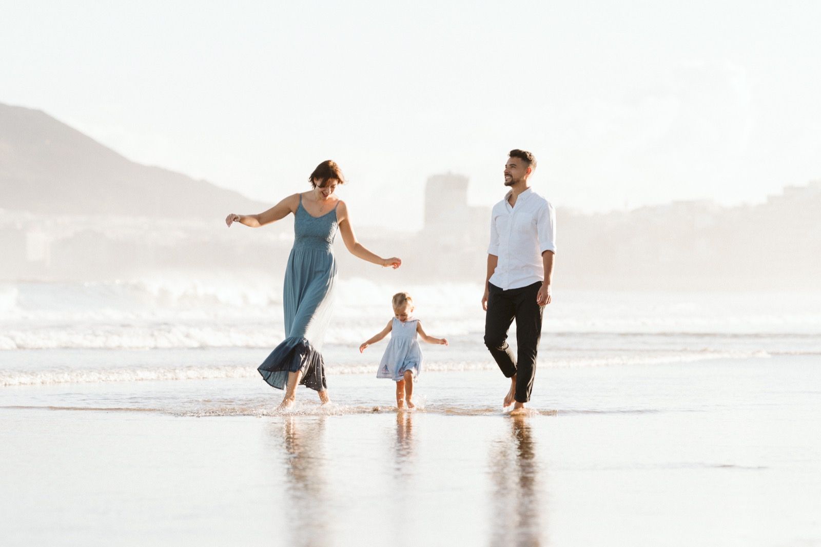Familia formada por hombre, mujer y niña pequeña posando a la cámara sonriendo y caminando por la orilla de la Playa de Las Canteras en Gran Canaria durante las horas de marea baja. Fotografía realizada por el fotógrafo de familias en Gran Canaria Héctor Cardeñosa.