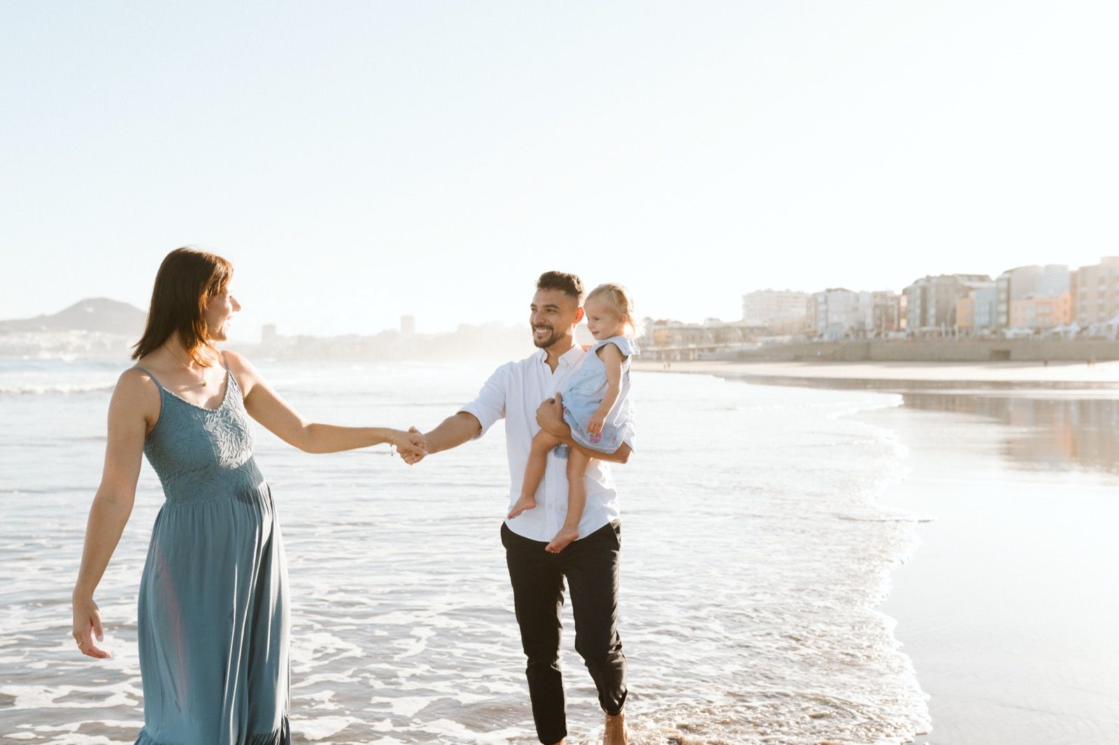 Familia formada por hombre, mujer y niña pequeña posando a la cámara sonriendo y caminando por la orilla de la Playa de Las Canteras en Gran Canaria durante las horas de marea baja. Fotografía realizada por el fotógrafo de familias en Gran Canaria Héctor Cardeñosa.