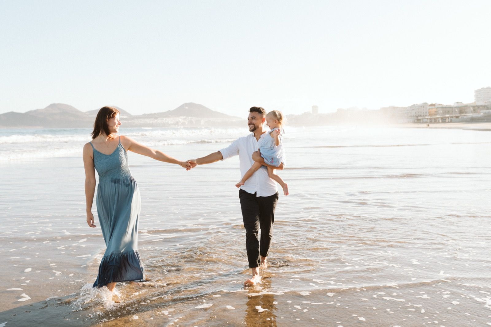Familia formada por hombre, mujer y niña pequeña posando a la cámara sonriendo y caminando por la orilla de la Playa de Las Canteras en Gran Canaria durante las horas de marea baja. Fotografía realizada por el fotógrafo de familias en Gran Canaria Héctor Cardeñosa.