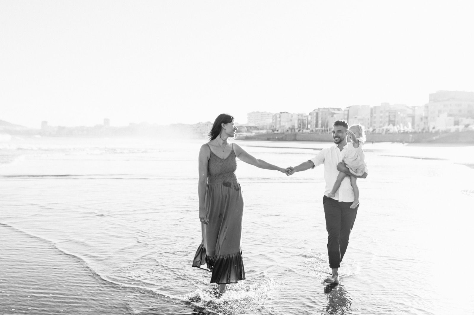 Familia formada por hombre, mujer y niña pequeña posando a la cámara sonriendo y caminando por la orilla de la Playa de Las Canteras en Gran Canaria durante las horas de marea baja. Fotografía realizada por el fotógrafo de familias en Gran Canaria Héctor Cardeñosa.