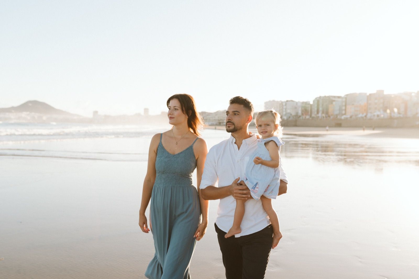 Familia formada por hombre, mujer y niña pequeña posando a la cámara sonriendo y caminando por la orilla de la Playa de Las Canteras en Gran Canaria durante las horas de marea baja. Fotografía realizada por el fotógrafo de familias en Gran Canaria Héctor Cardeñosa.