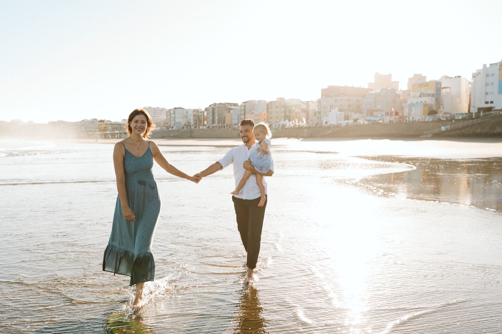 Familia formada por hombre, mujer y niña pequeña posando a la cámara sonriendo y caminando por la orilla de la Playa de Las Canteras en Gran Canaria durante las horas de marea baja. Fotografía realizada por el fotógrafo de familias en Gran Canaria Héctor Cardeñosa.