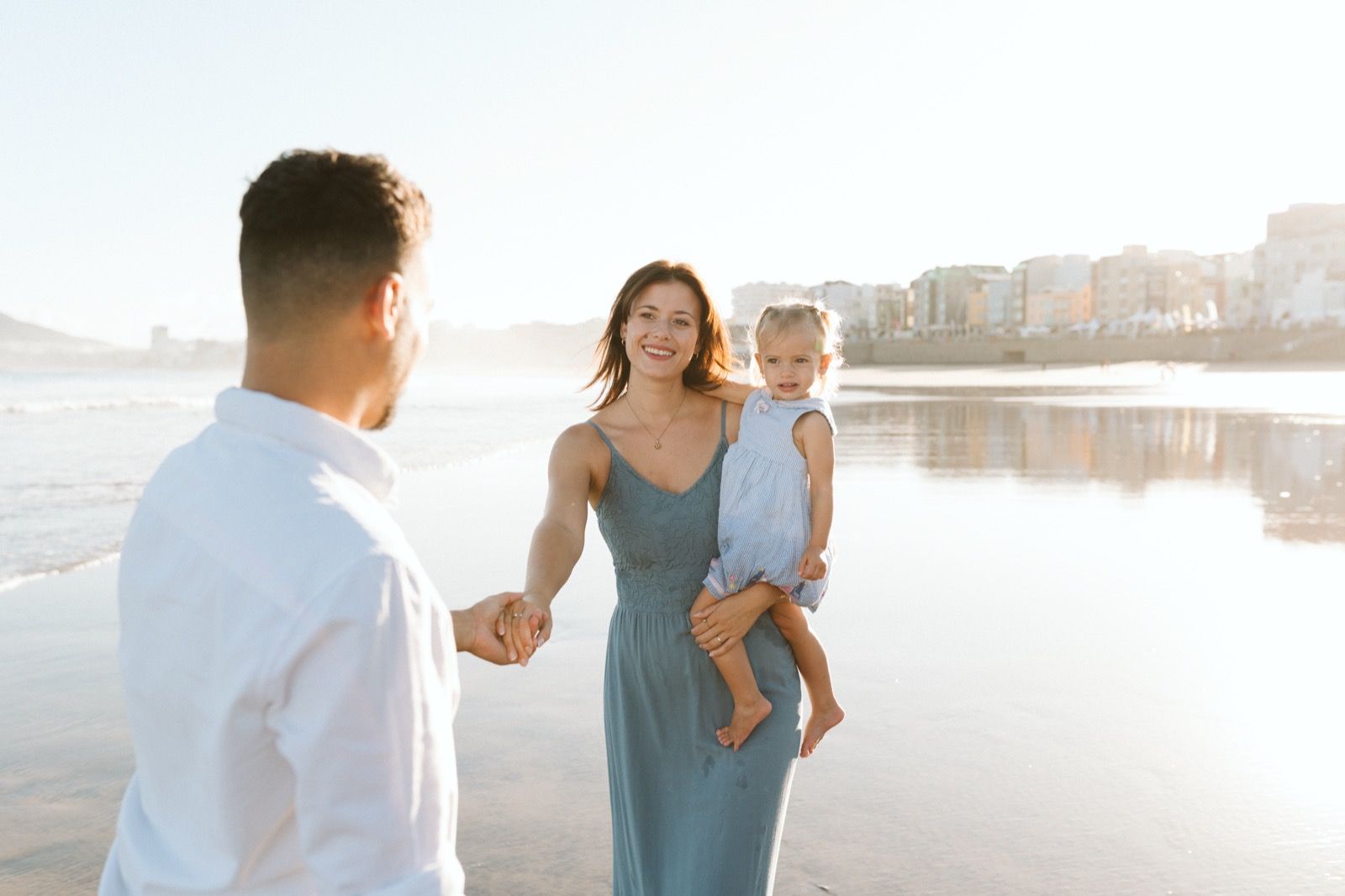 Familia formada por hombre, mujer y niña pequeña posando a la cámara sonriendo y caminando por la orilla de la Playa de Las Canteras en Gran Canaria durante las horas de marea baja. Fotografía realizada por el fotógrafo de familias en Gran Canaria Héctor Cardeñosa.