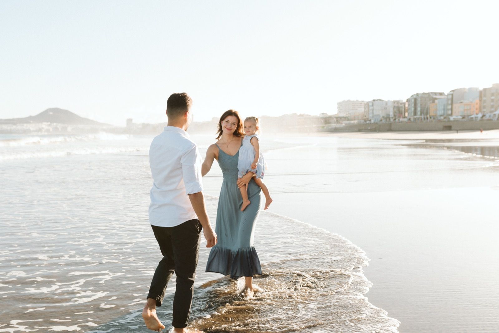 Familia formada por hombre, mujer y niña pequeña posando a la cámara sonriendo y caminando por la orilla de la Playa de Las Canteras en Gran Canaria durante las horas de marea baja. Fotografía realizada por el fotógrafo de familias en Gran Canaria Héctor Cardeñosa.