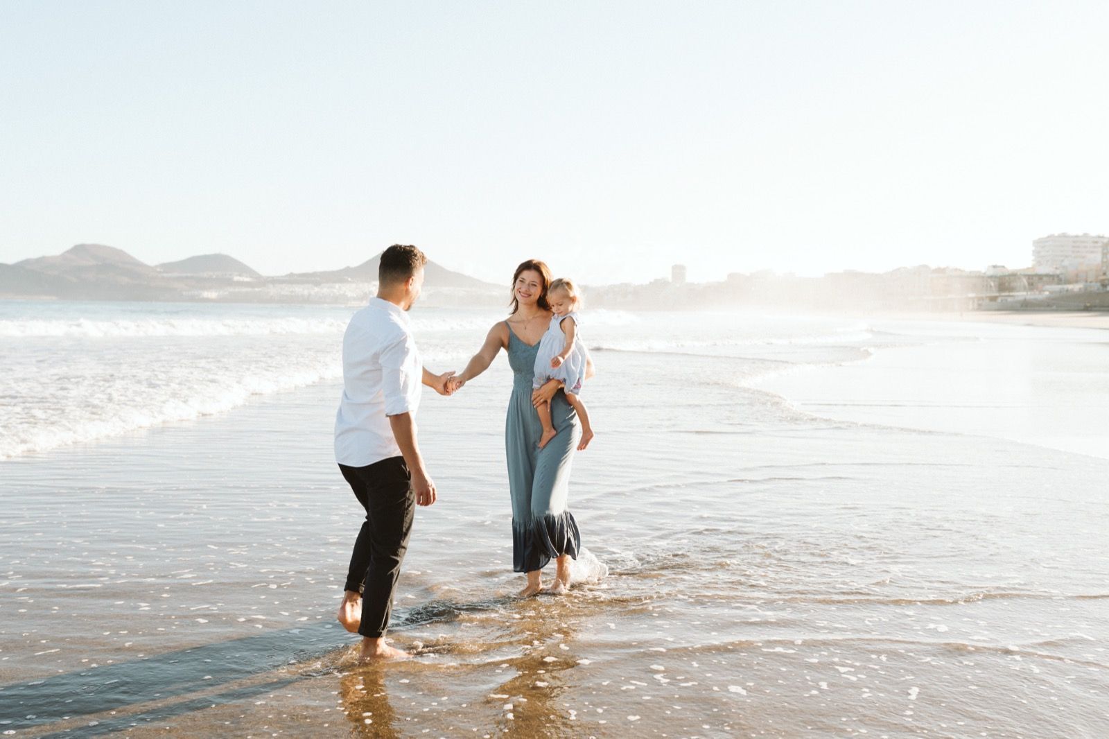 Familia formada por hombre, mujer y niña pequeña posando a la cámara sonriendo y caminando por la orilla de la Playa de Las Canteras en Gran Canaria durante las horas de marea baja. Fotografía realizada por el fotógrafo de familias en Gran Canaria Héctor Cardeñosa.