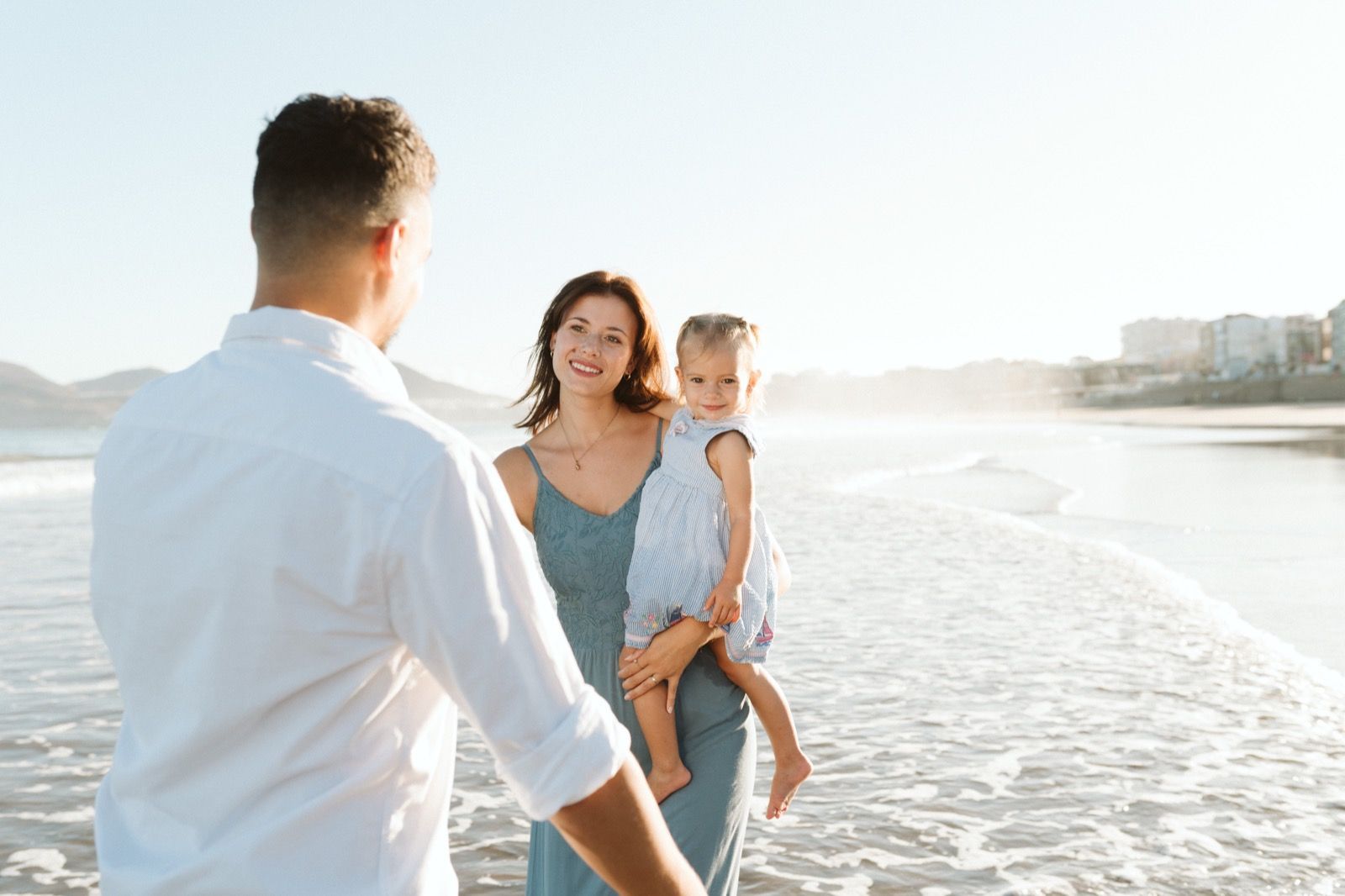 Familia formada por hombre, mujer y niña pequeña posando a la cámara sonriendo en la orilla de la Playa de Las Canteras en Gran Canaria durante las horas de marea baja. Fotografía realizada por el fotógrafo de familias en Gran Canaria Héctor Cardeñosa.
