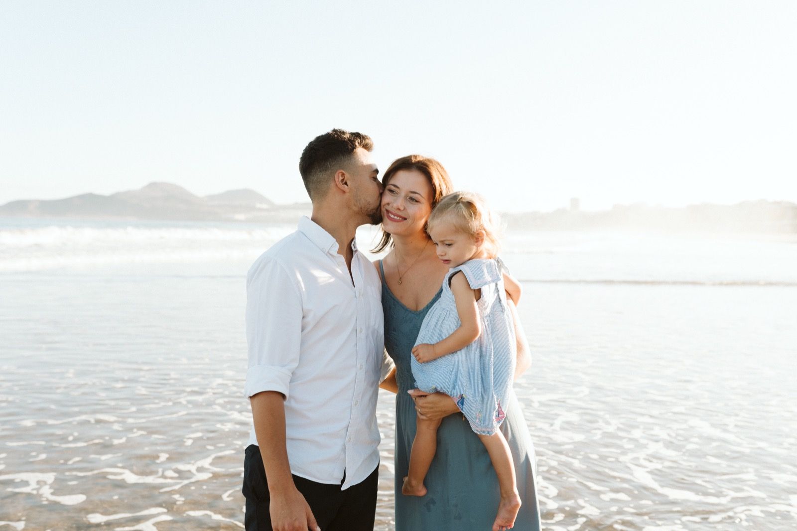 Familia formada por hombre, mujer y niña pequeña posando a la cámara sonriendo en la orilla de la Playa de Las Canteras en Gran Canaria durante las horas de marea baja. Fotografía realizada por el fotógrafo de familias en Gran Canaria Héctor Cardeñosa.