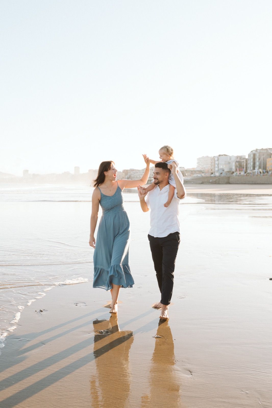 Familia formada por hombre, mujer y niña pequeña posando a la cámara sonriendo y caminando por la orilla de la Playa de Las Canteras en Gran Canaria durante las horas de marea baja. Fotografía realizada por el fotógrafo de familias en Gran Canaria Héctor Cardeñosa.