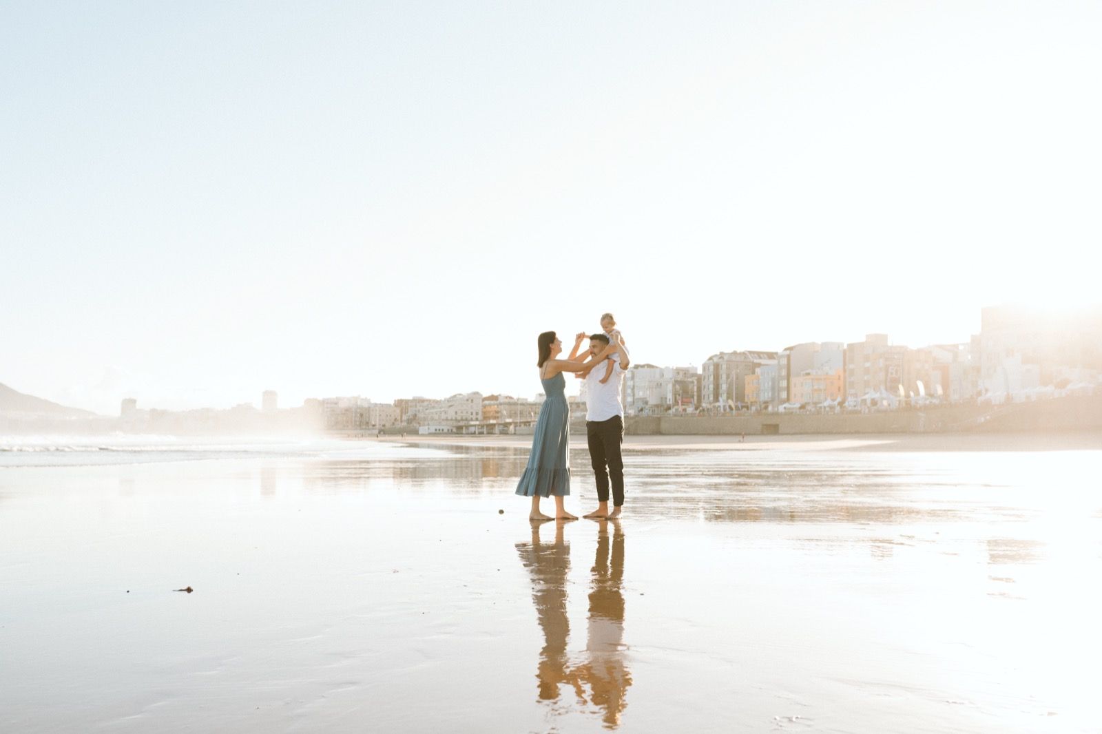 Familia formada por hombre, mujer y niña pequeña posando a la cámara sonriendo y caminando por la orilla de la Playa de Las Canteras en Gran Canaria durante las horas de marea baja. Fotografía realizada por el fotógrafo de familias en Gran Canaria Héctor Cardeñosa.