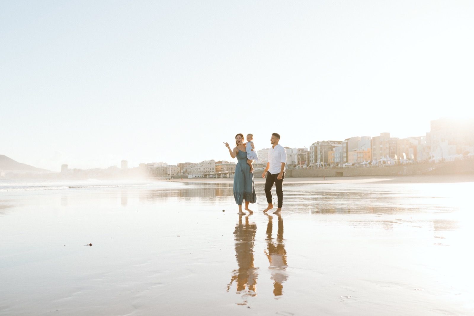Familia formada por hombre, mujer y niña pequeña posando a la cámara sonriendo y caminando por la orilla de la Playa de Las Canteras en Gran Canaria durante las horas de marea baja. Fotografía realizada por el fotógrafo de familias en Gran Canaria Héctor Cardeñosa.