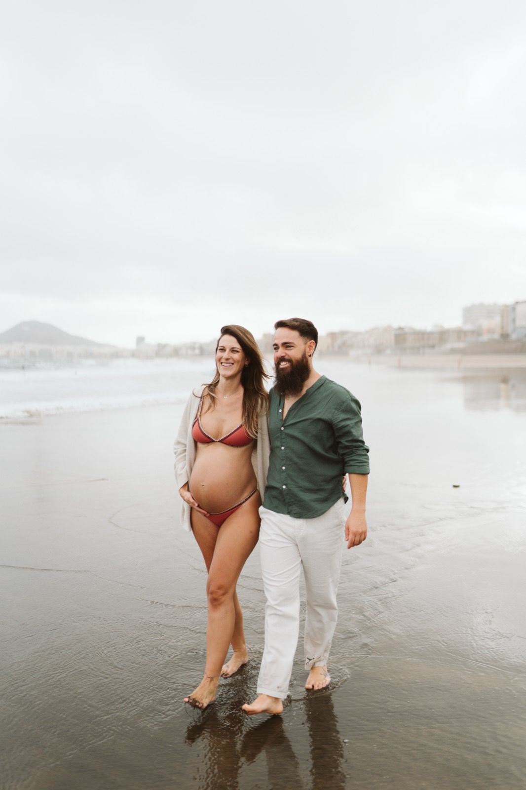 Pareja caminando sonrientes por la orilla de la playa durante unas fotos de embarazo en la Playa de Las Canteras