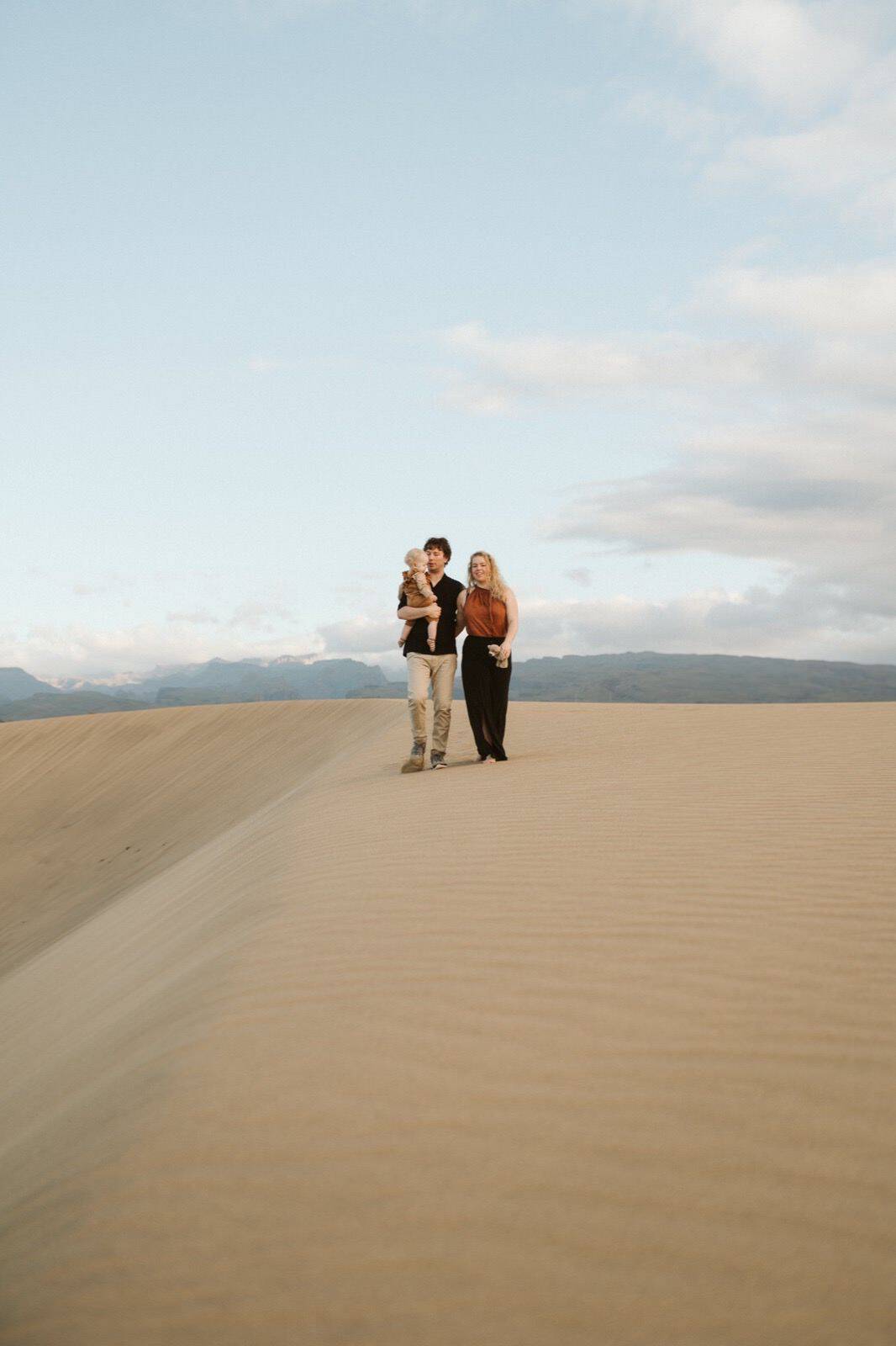 Pareja mirándose y caminando con su hijo en brazos en una sesión de fotos familiares en Gran Canaria, rodeados de naturaleza en las Dunas de Maspalomas