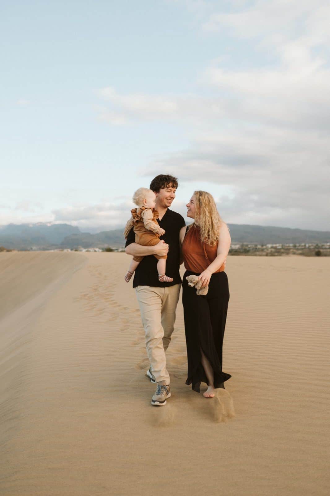 Pareja mirándose y caminando con su hijo en brazos en una sesión de fotos familiares en Gran Canaria, rodeados de naturaleza en las Dunas de Maspalomas